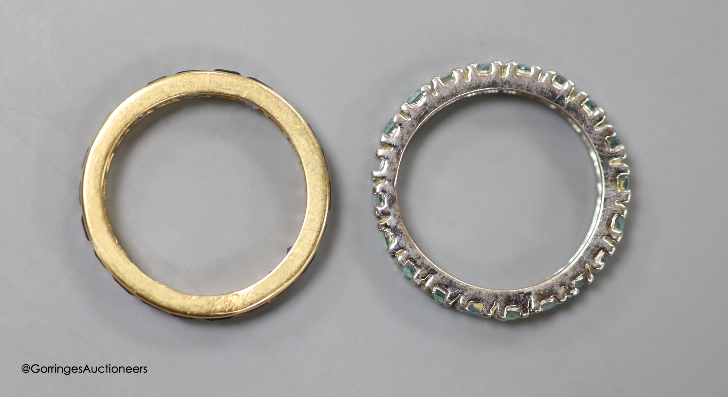 A yellow metal and sapphire set full eternity ring, size M, gross weight 2.8 grams and a white metal and blue gem set full eternity ring, size N/O, gross 2.4 grams.